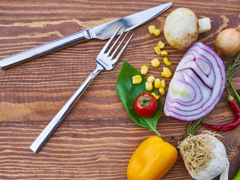 A knife and fork sit next to a selection of vegetables including an onion, yellow pepper, mushroom, and garlic, all part of a vegan diet.