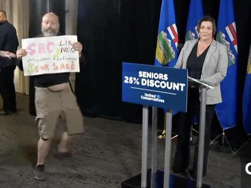 A protester waves a sign for a a few seconds at the United Conservative Party’s news conference yesterday while Premier Danielle Smith stands calmly nearby, obviously in no danger.