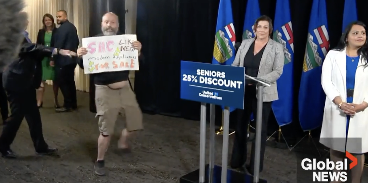 A protester waves a sign for a a few seconds at the United Conservative Party’s news conference yesterday while Premier Danielle Smith stands calmly nearby, obviously in no danger.