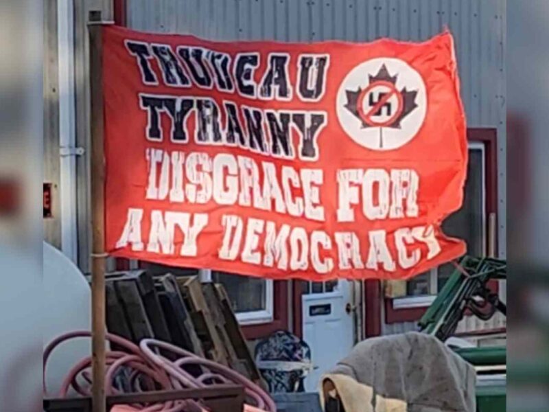 A read flag with the words Trudeau Tyranny a Disgrace for Any Democracy written on it with a maple leave with a Nazi swastika over it with a red line through the symbol.
