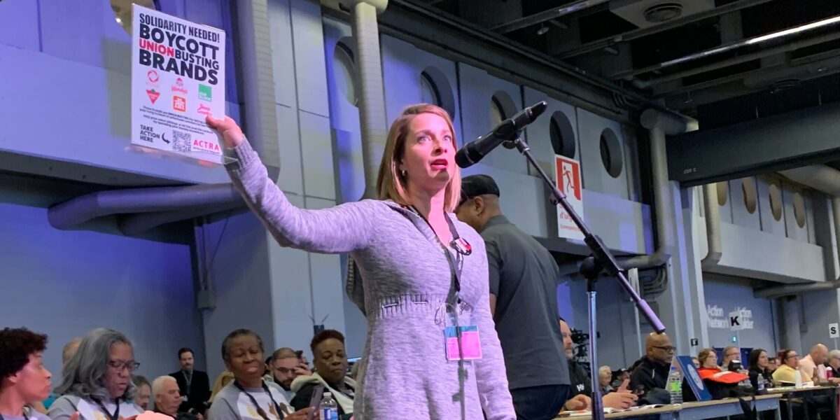 Eleanor Noble, National President of ACTRA stands in front of a microphone holding up a poster calling for boycotts of Canadian brand names that are locking out her members.