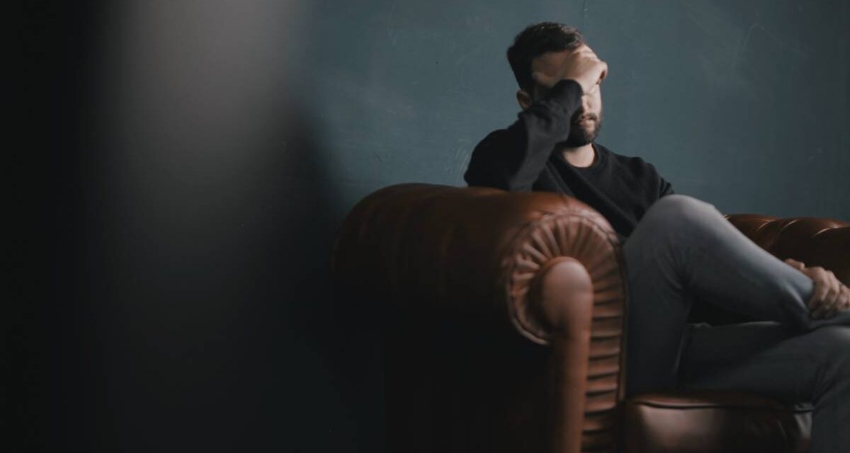 A man sitting on a couch with a hand covering his face.