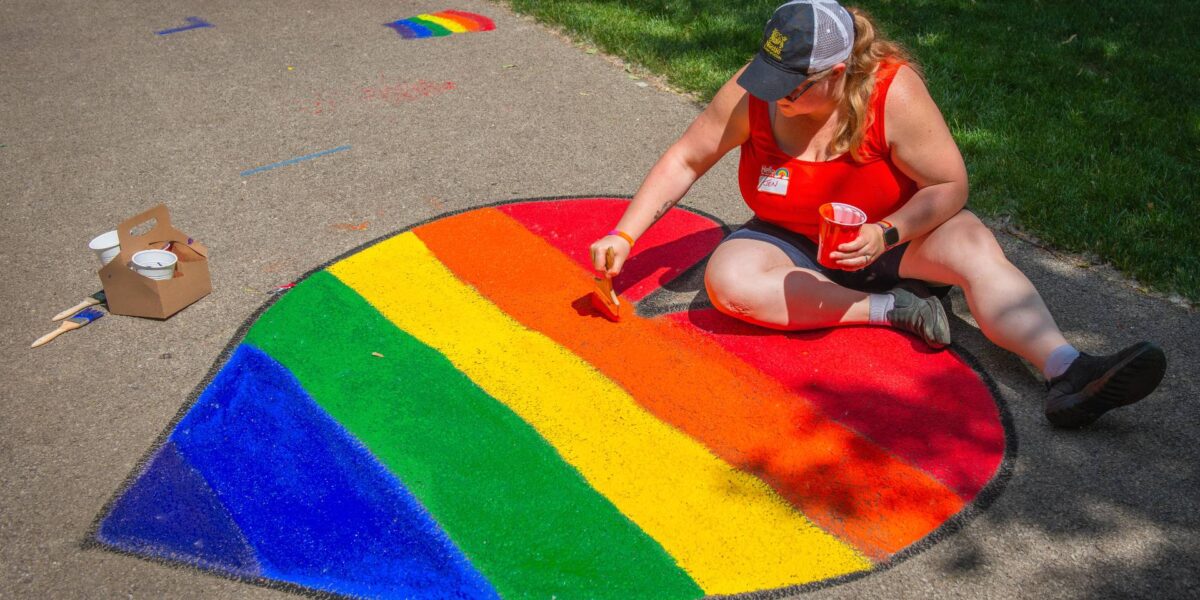 A person in a baseball cap draws a pride flag in chalk in the shape of a heart on sidewalk pavement.