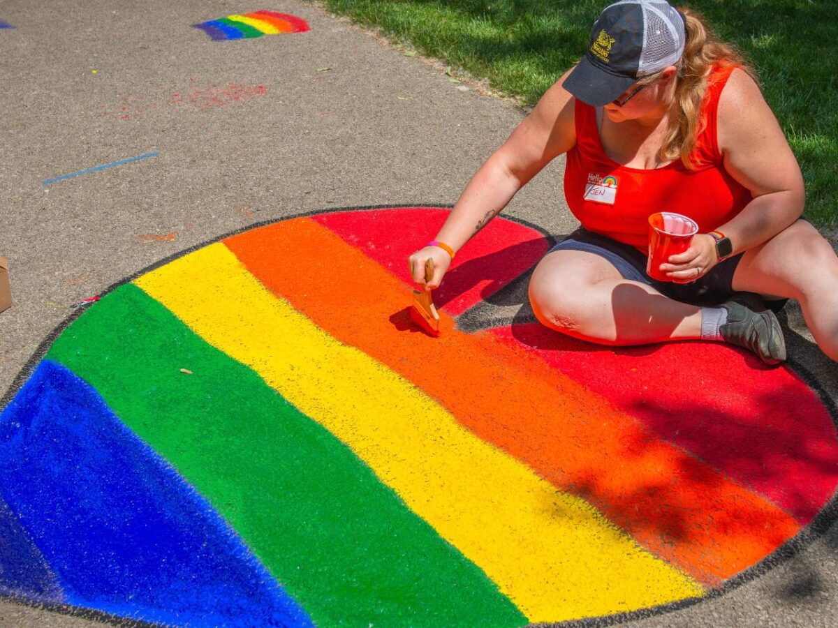 A person in a baseball cap draws a pride flag in chalk in the shape of a heart on sidewalk pavement.