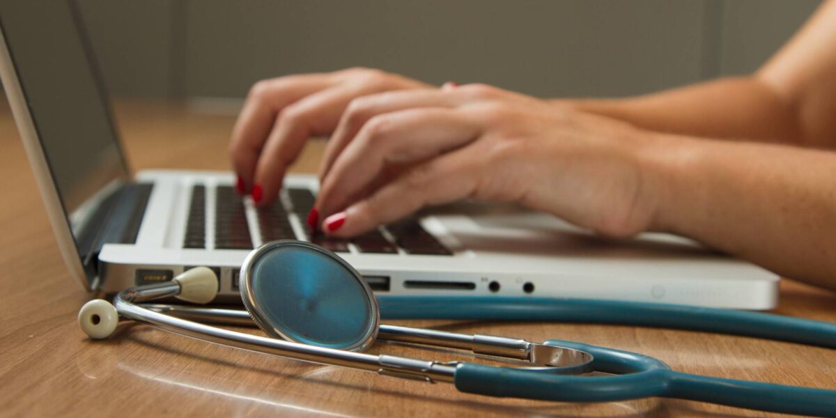 A physician typing on a laptop with a stethoscope in the foreground.