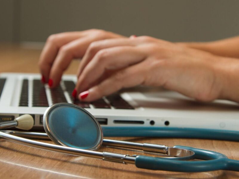 A physician typing on a laptop with a stethoscope in the foreground.