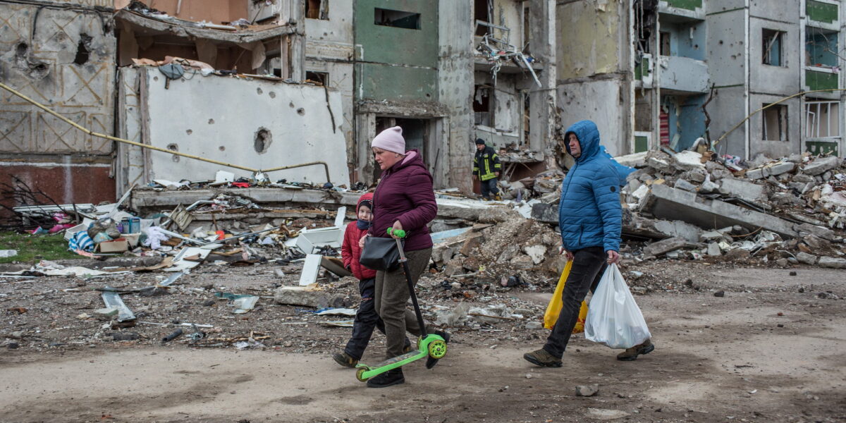 People walking down street along crumbling buildings in Ukraine.