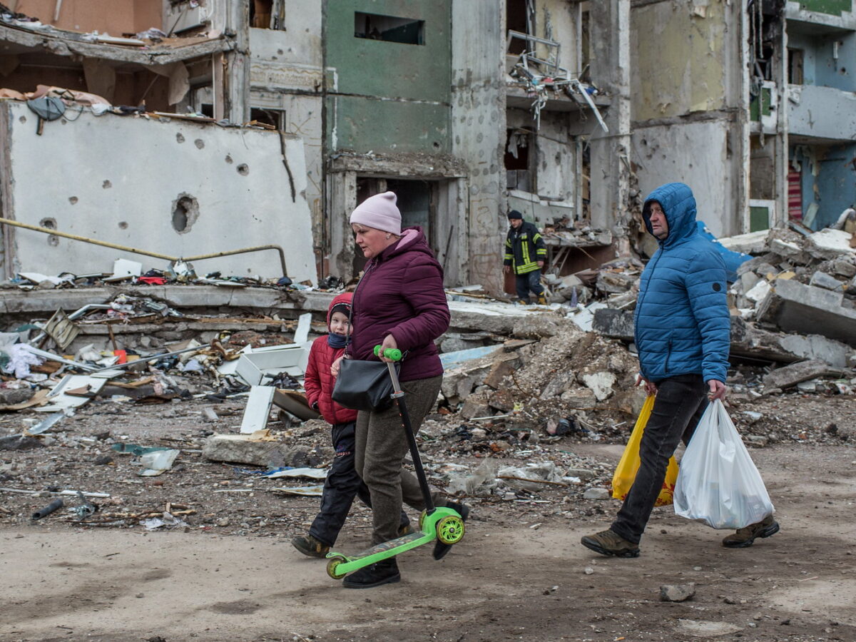 People walking down street along crumbling buildings in Ukraine.