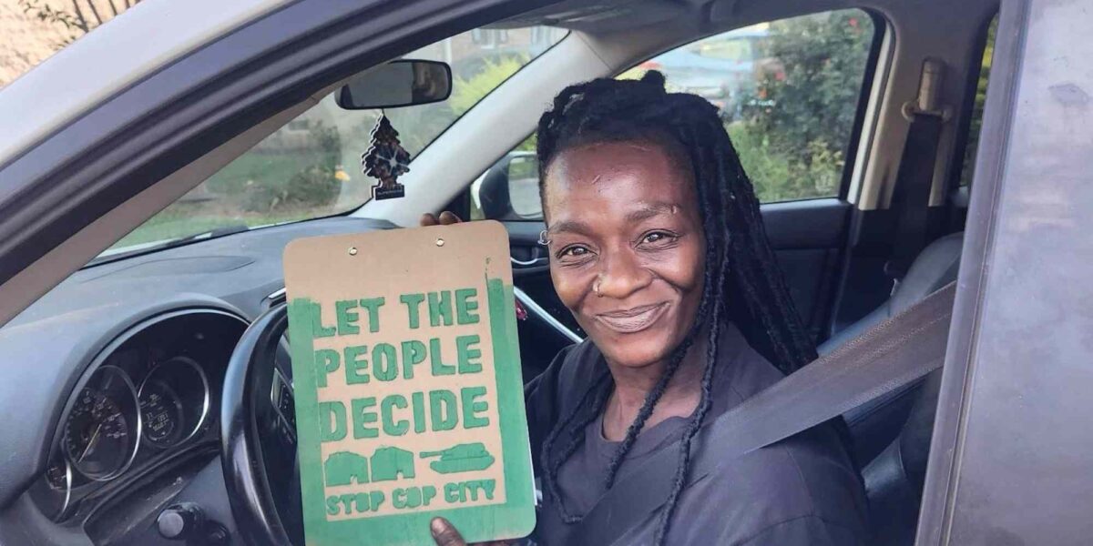 A Black woman sits in a car holding a sign encouraging citizens to vote against Cop City.