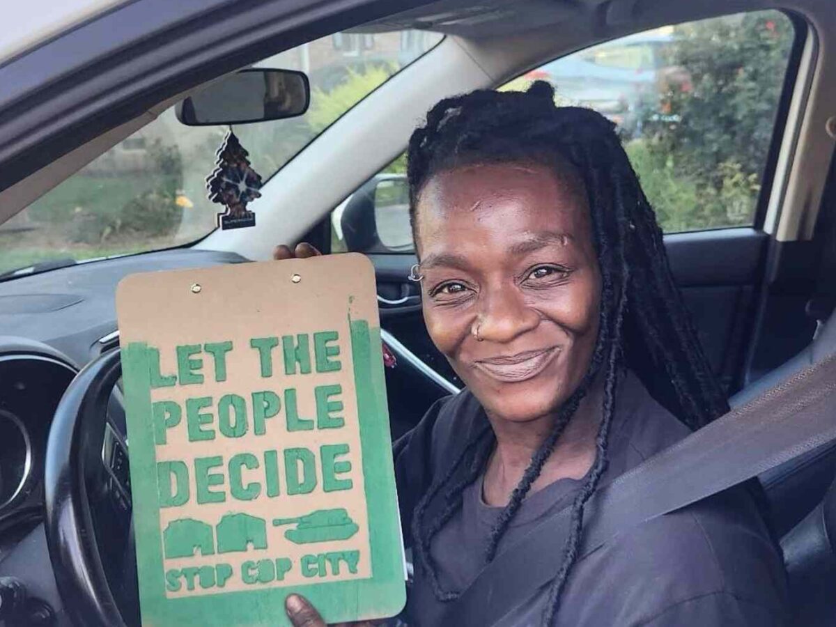 A Black woman sits in a car holding a sign encouraging citizens to vote against Cop City.