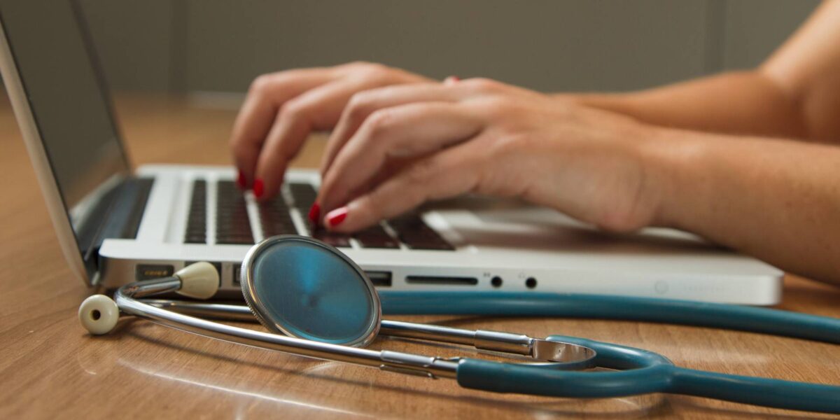 A pair of hands types on a laptop with a stethoscope in the foreground.