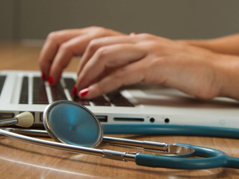 A pair of hands types on a laptop with a stethoscope in the foreground.