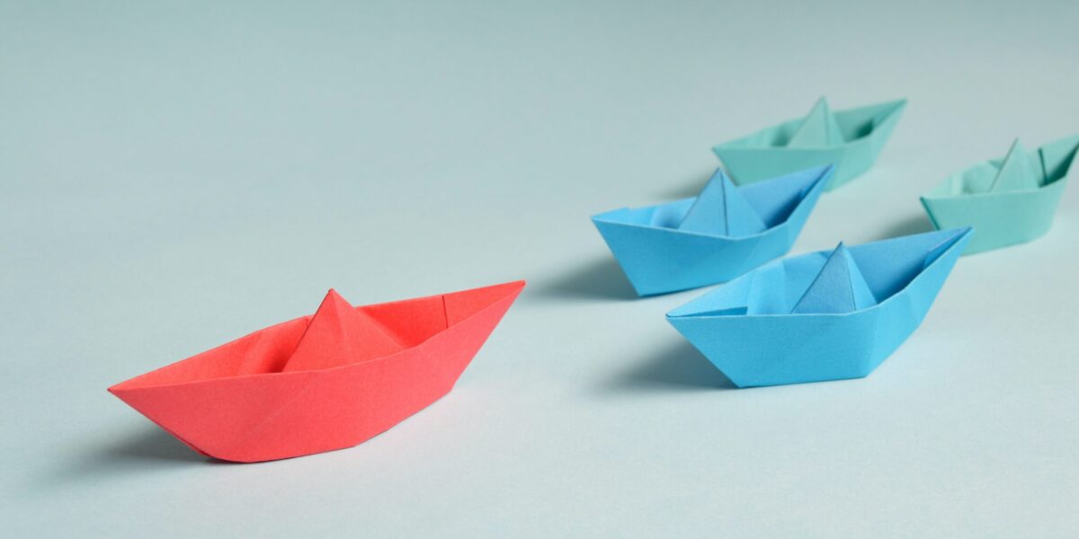 4 blue and green paper boats being led by a red one