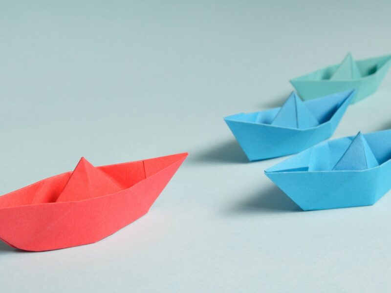 4 blue and green paper boats being led by a red one