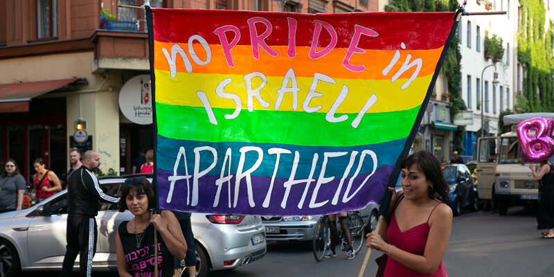 Two women hold a rainbow Pride flag with the words "No Pride in Israel Apartheid" on it.