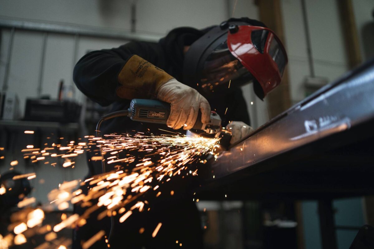 A person wears a welding mask as they work on a piece of metal. Sparks fly.