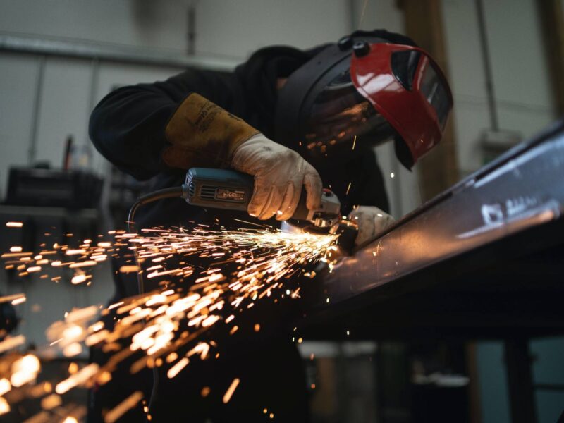 A person wears a welding mask as they work on a piece of metal. Sparks fly.