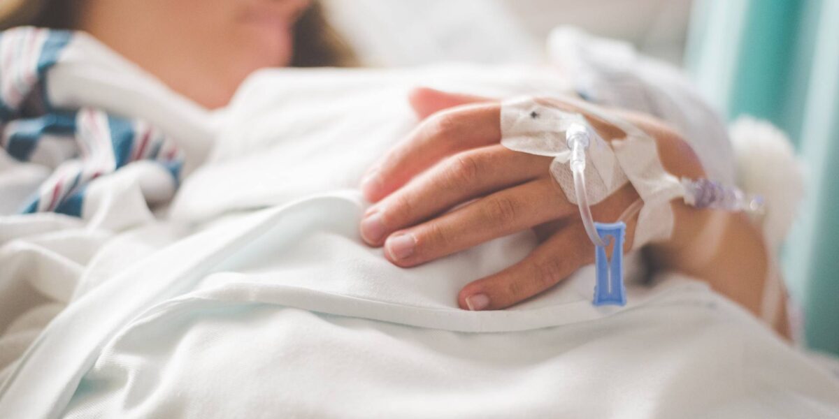 A person receiving hospital care lying on a bed with an intravenous tube in their hand.