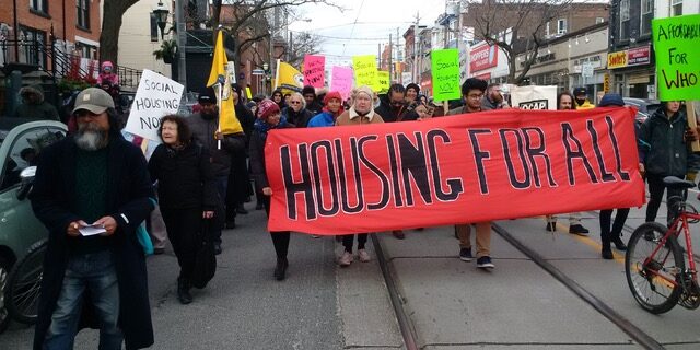 Protesters march during a National Housing Day protest in 2017 with individuals at the front of the march holding a red banner with the words: "Housing for all" written on it.