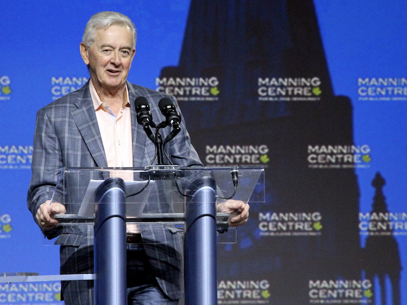 Preston Manning standing at a glass podium in front of a blue background.