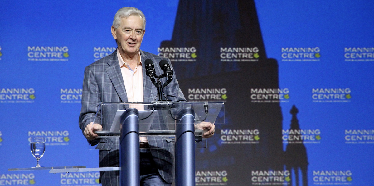 Preston Manning standing at a glass podium in front of a blue background.