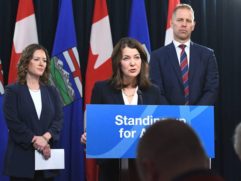 Alberta Environment Minister Rebecca Schulz and Utilities Minister Nathan Neudorf look on as Premier Danielle Smith speaks from a podium at a press conference.