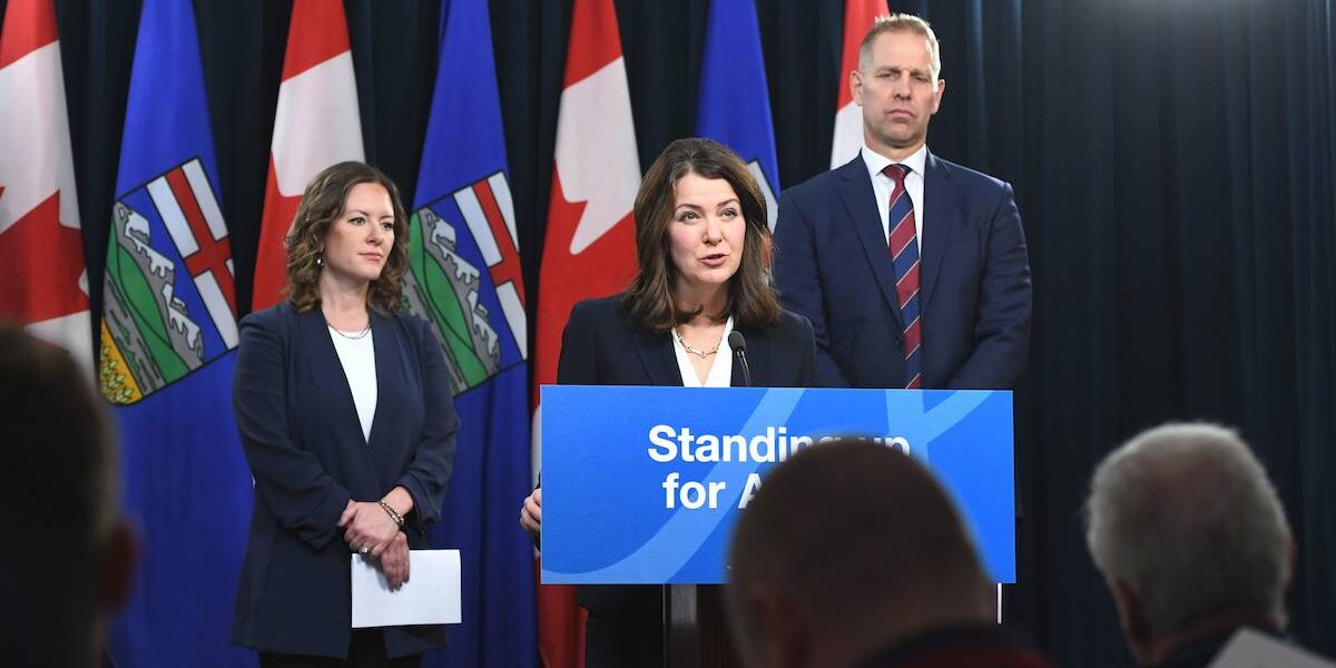 Alberta Environment Minister Rebecca Schulz and Utilities Minister Nathan Neudorf look on as Premier Danielle Smith speaks from a podium at a press conference.