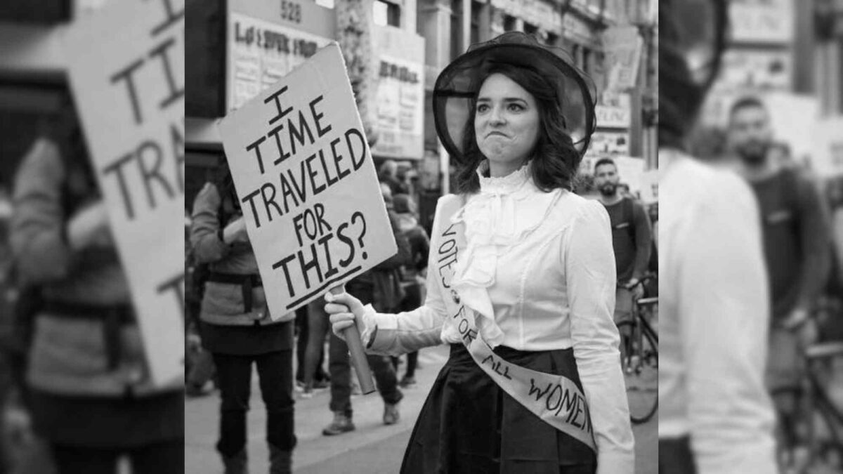 A black and white photo of a woman in 19th century dress holding a sign that reads "I time travelled for this."