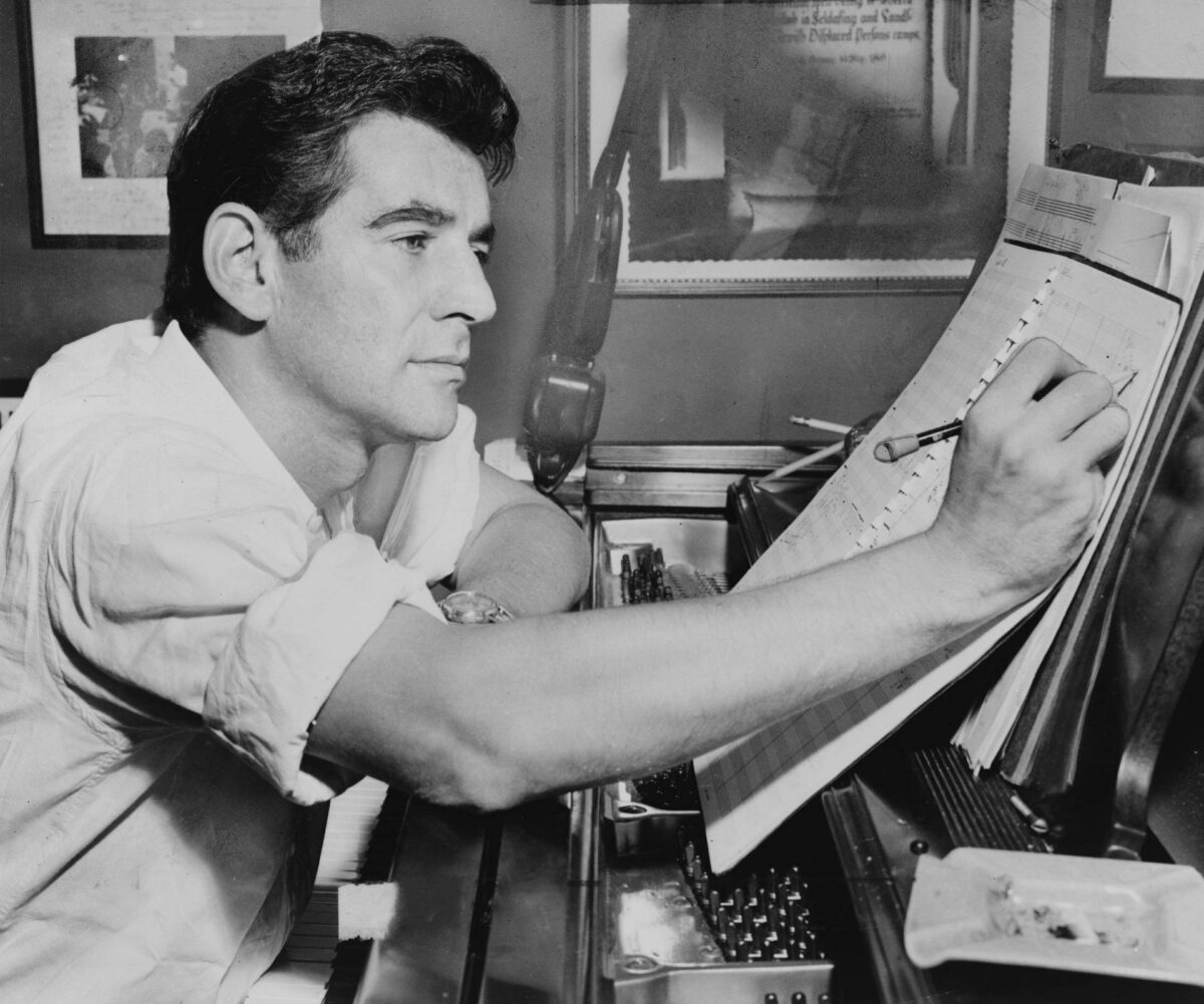 Leonard Bernstein seated at piano, making annotations to musical score.