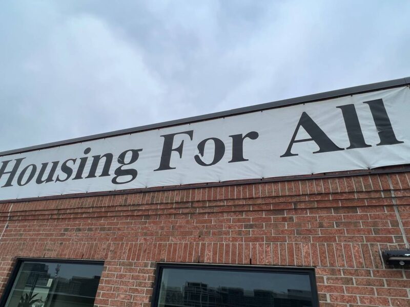 A Housing For All banner on a Toronto housing co-op with the one per cent logo from the 1998 campaign for all levels of government to allocate an additional one per cent of their budget to a national housing program.