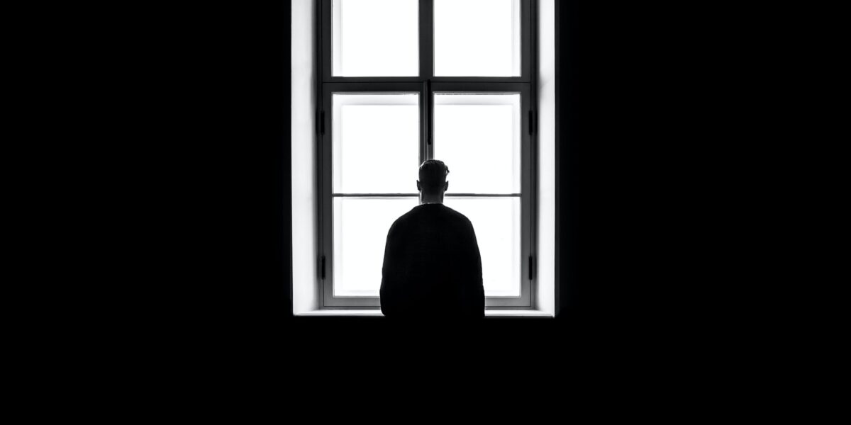 A black and white photo of a single person looking out a window with their back to the camera.