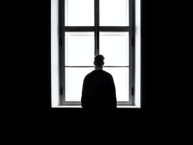 A black and white photo of a single person looking out a window with their back to the camera.