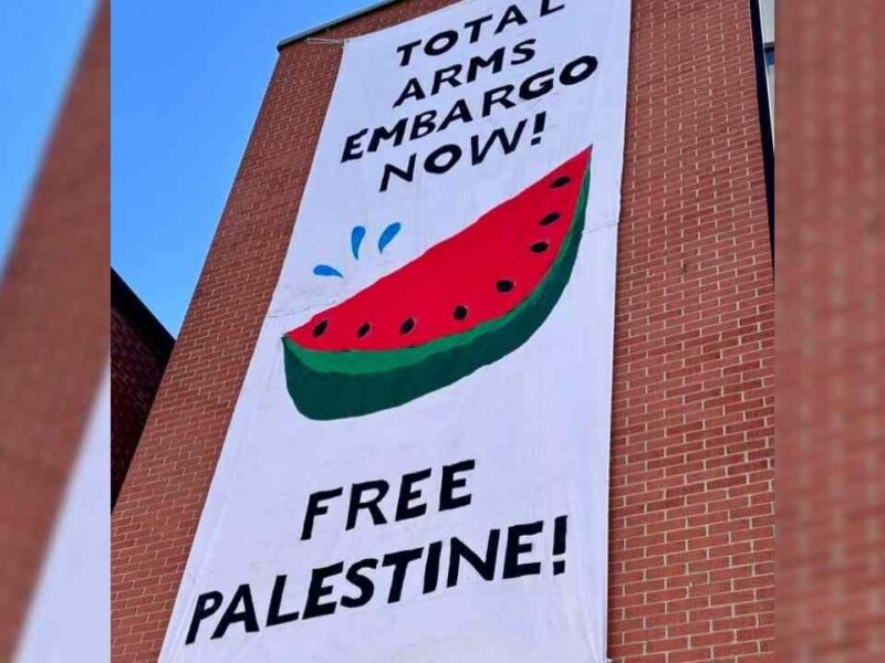 A white banner with a watermelon slice on it hangs from a building in down town Ottawa and reads "Total arms embargo now! Free Palestine."