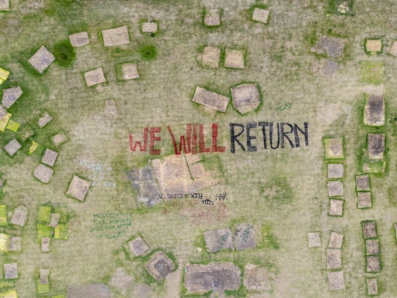 An aerial photo of the vacated encampment site with the words "We Will Return" painted on the earth.