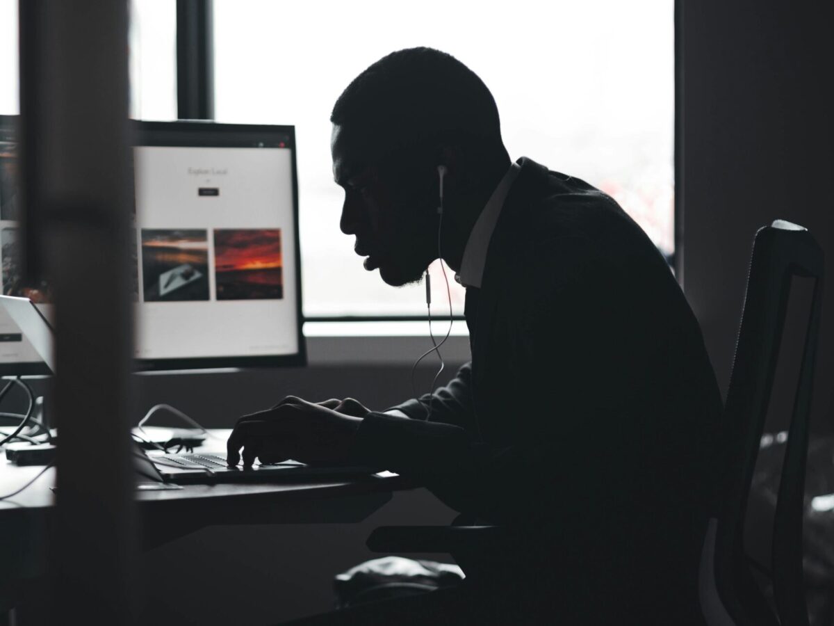 A silhouetted man is hunched over a laptop. Behind him, another computer monitor is running.