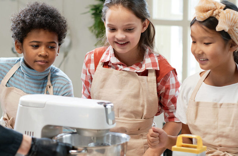 An image of three kids watching an adult demonstrate how to use a stand mixer.