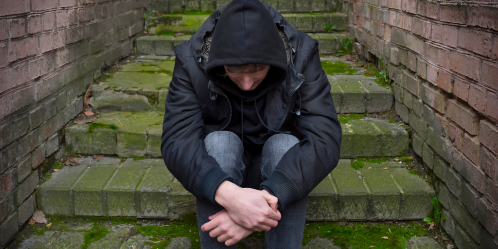 A white man sitting on steps outside, wearing a hoodie and looking down.