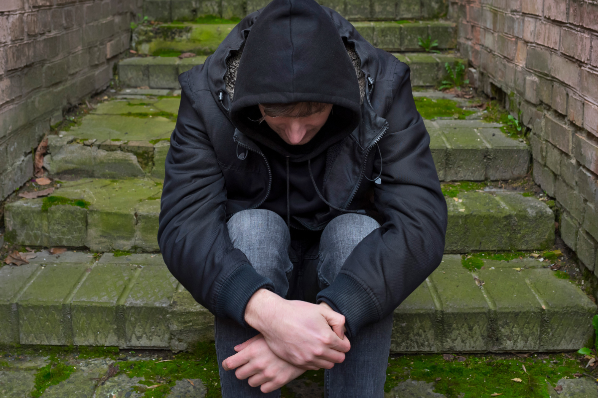 A white man sitting on steps outside, wearing a hoodie and looking down.