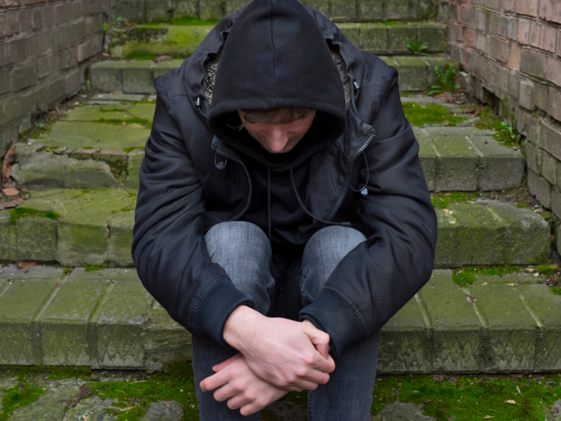 A white man sitting on steps outside, wearing a hoodie and looking down.