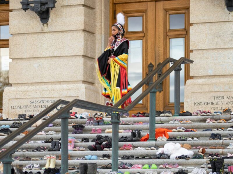 An image of a woman sin traditional clothing standing at the top of the steps of a building covered in children's shoes. Acknowledging the scars of residential schools is part of truth and reconciliation.