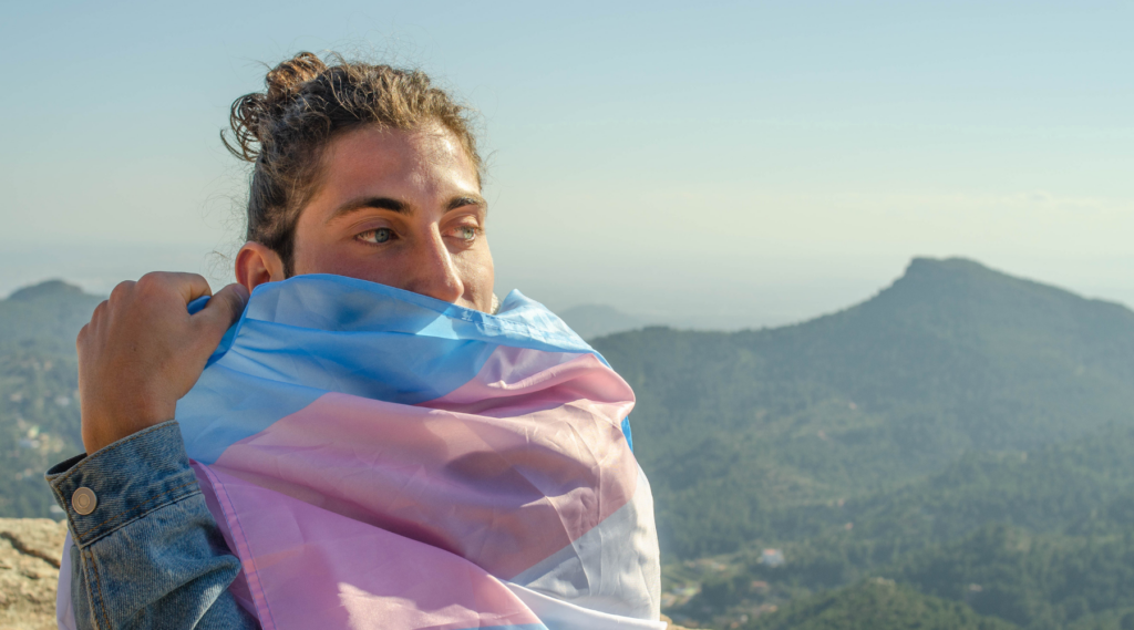 Person wrapping a trans pride flag around themselves on top of a mountain.