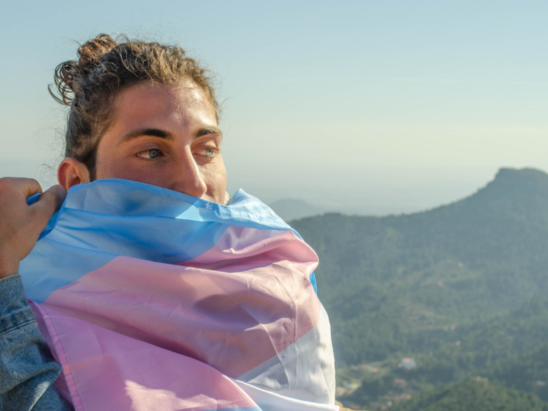 Person wrapping a trans pride flag around themselves on top of a mountain.