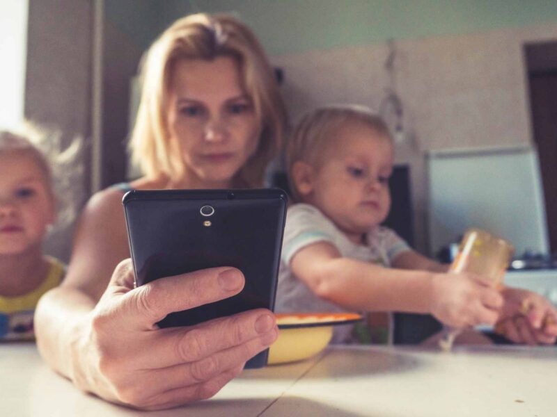 A woman cares for two children while checking emails on her phone.