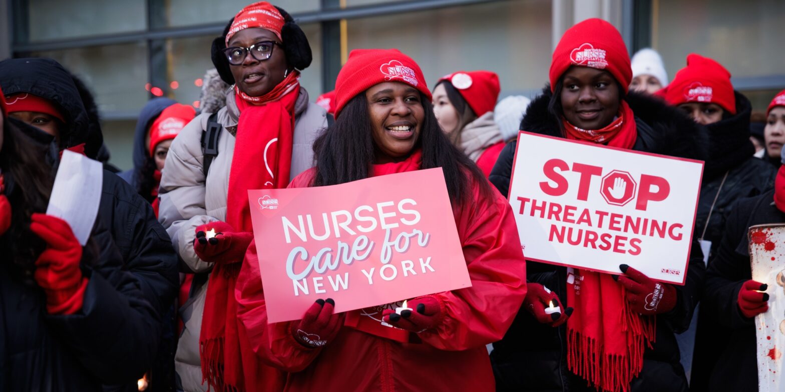 An image of New York City nurses on the picket line. Two nurses wearing red winter hats and red coats are in focus. The nurse on the left is holding a sign that says, "Nurses care for New York." The nurse on the right holds a sign that says, "Stop threatening nurses."