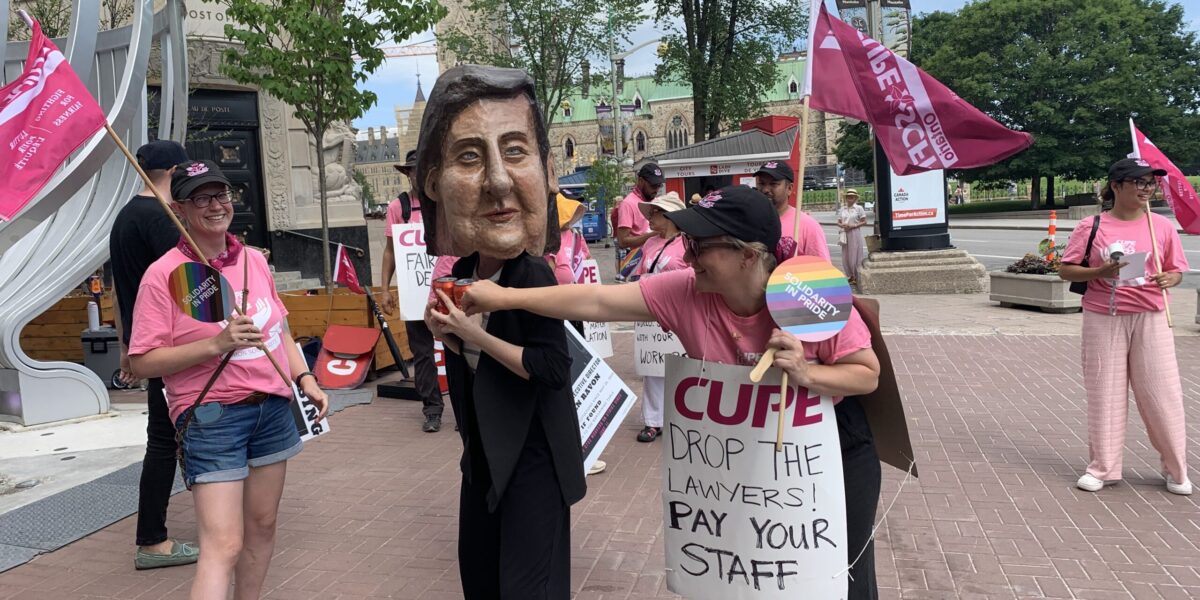 A striking worker wearing a giant Lauren Ravon head hoard cans of cola, representing cost-of-living-adjustments, on the Ottawa picket line July 8, 2026.