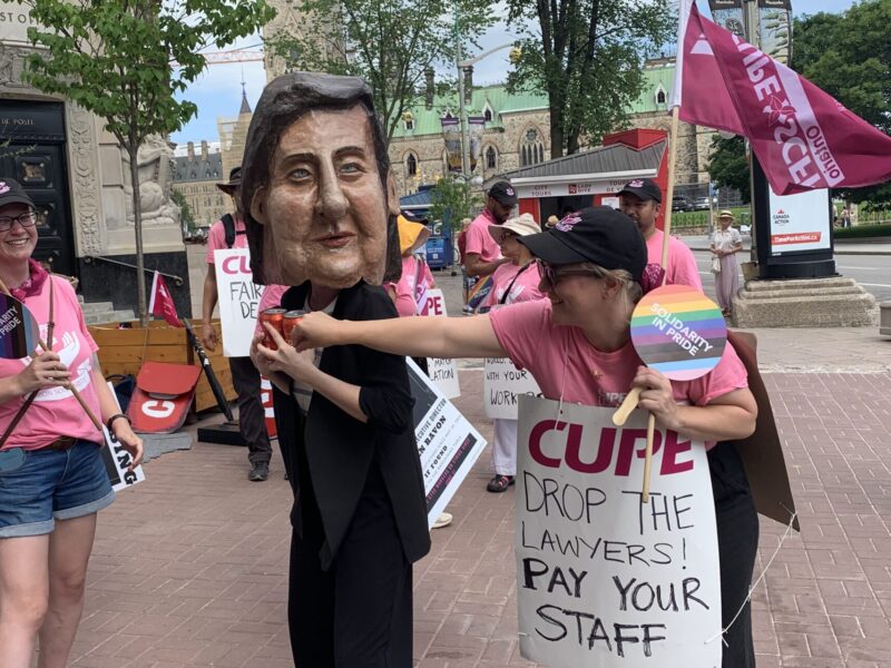 A striking worker wearing a giant Lauren Ravon head hoard cans of cola, representing cost-of-living-adjustments, on the Ottawa picket line July 8, 2026.